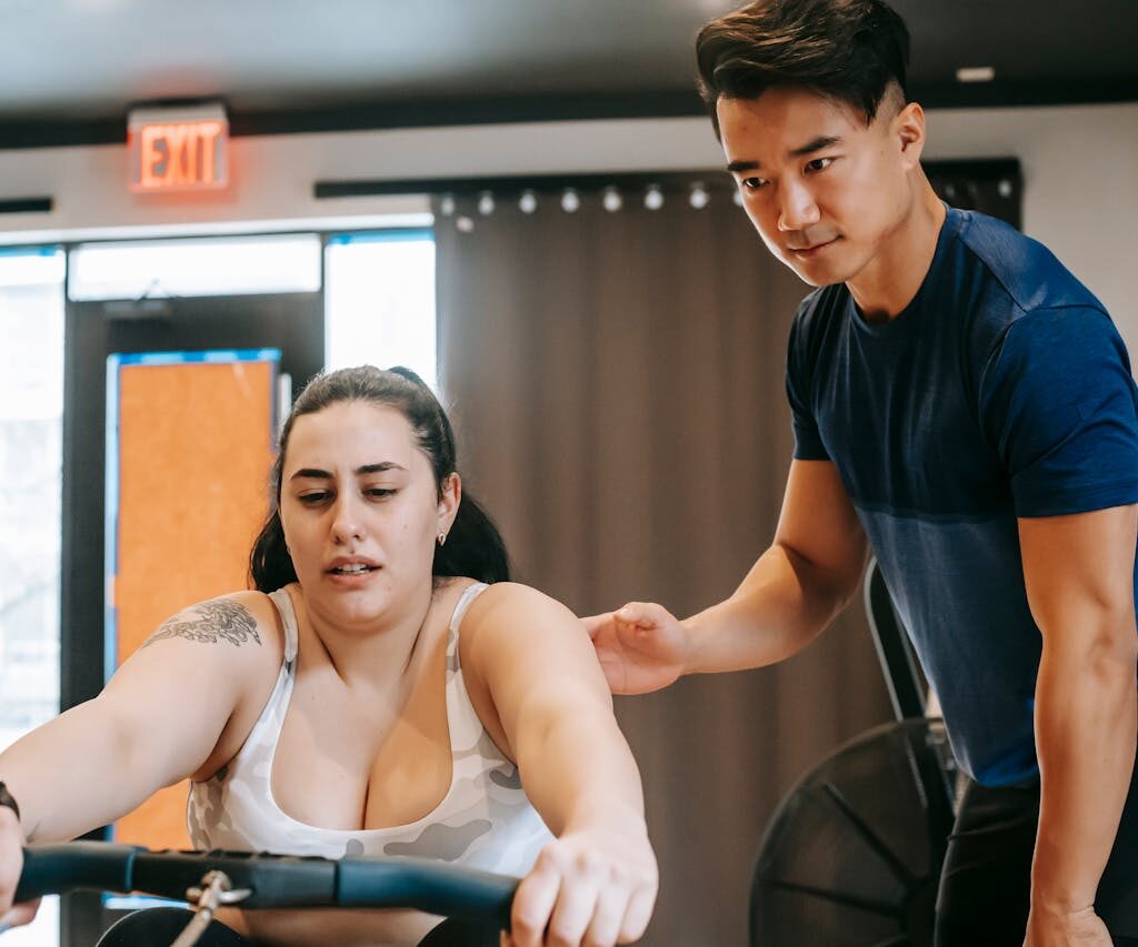 Young handsome ethnic experienced trainer supporting plus size woman working out on rowing machine in gym